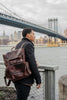 Person wearing a brown leather backpack with a cityscape and bridge in the background